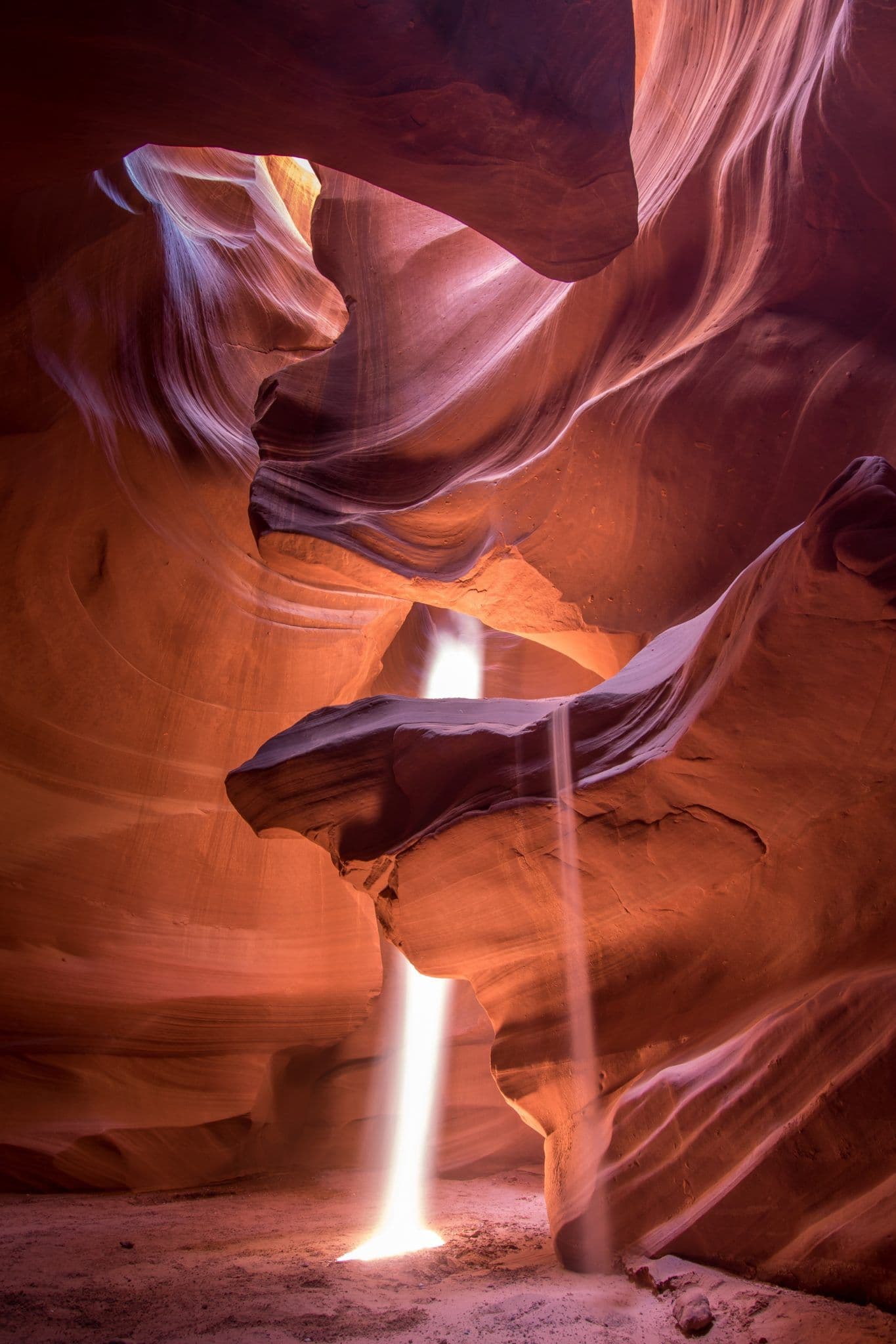 This image captures a breathtaking natural rock formation within a narrow, winding canyon, characterized by smooth, undulating sandstone walls that swirl in warm hues of orange, red, and deep purple. A vertical beam of sunlight pierces through an opening above, illuminating the cavern floor and casting dramatic, elongated light streaks that contrast with the shadowed, textured rock surfaces. The composition emphasizes the organic, sculptural curves of the canyon, suggesting geological erosion over millennia, while the interplay of light and shadow creates a sense of depth and mystery. The scene is rendered with high photographic precision, showcasing the natural beauty and serene grandeur of the landscape, evoking a tranquil, awe-inspiring atmosphere. The absence of human presence or artificial elements enhances the image's focus on natural wonder and the sublime power of nature.