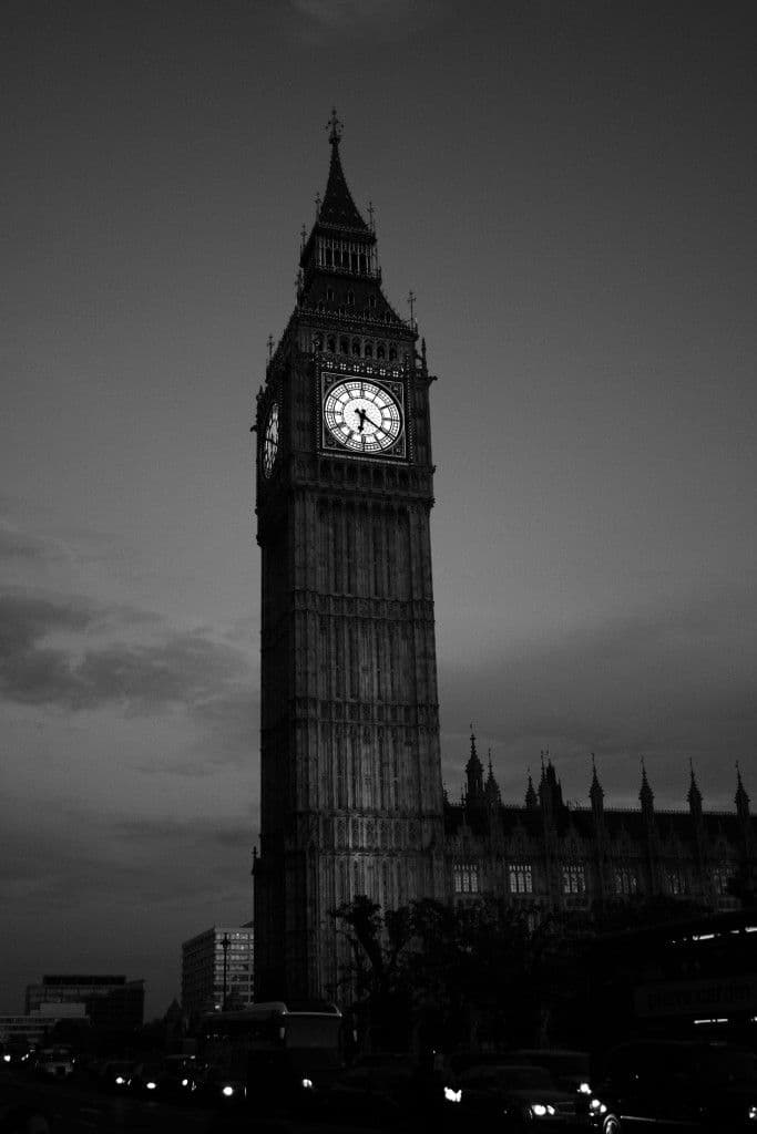 This black-and-white photograph captures the iconic Elizabeth Tower, commonly known as Big Ben, standing tall against a dusky sky. The clock face is illuminated, contrasting sharply with the darkened stone architecture and surrounding buildings. Below, the silhouettes of vehicles and urban structures suggest a bustling cityscape at twilight. The image employs dramatic chiaroscuro lighting, emphasizing the tower's verticality and architectural detail. The overall atmosphere is solemn and majestic, evoking a sense of historical permanence and urban grandeur. The composition is a medium shot, centered on the tower, with a shallow depth of field that blurs the foreground vehicles, drawing focus to the monument's imposing presence.