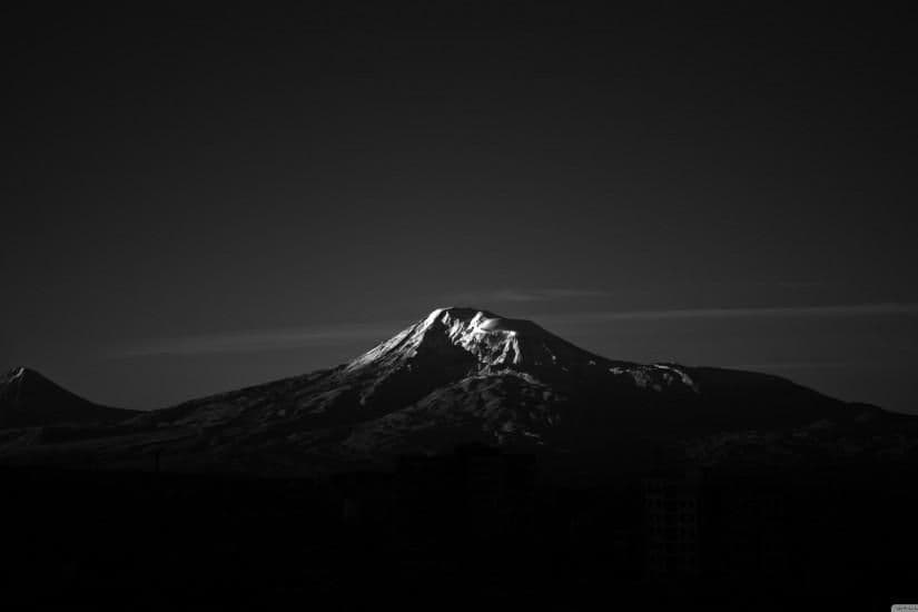 This is a high-contrast black and white photograph capturing a majestic, snow-capped mountain peak silhouetted against a dark, clear sky. The mountain's rugged contours are sharply defined, with the snow reflecting light on its summit, creating a dramatic focal point. In the foreground, the dark silhouettes of urban buildings suggest a cityscape nestled at the base of the mountain, adding scale and context. The composition emphasizes the grandeur of nature juxtaposed with human habitation. The image employs minimalist tonal contrast and a stark, cinematic aesthetic to evoke a sense of awe and solitude. The overall mood is serene and contemplative, inviting quiet reflection on the vastness of the natural world.