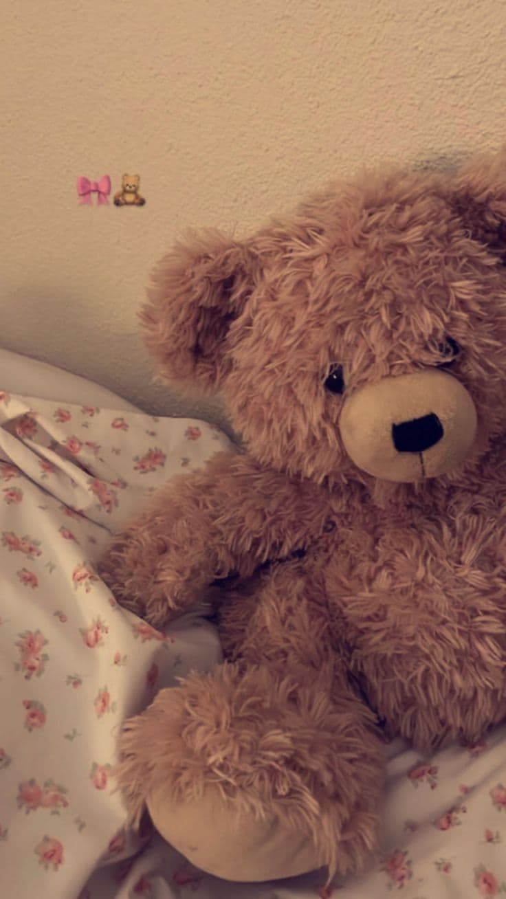 A plush, medium-brown teddy bear with soft, shaggy fur sits comfortably on a bed covered in a white sheet featuring small pink floral patterns. The bear's facial features are simple and endearing, with black button eyes, a stitched nose, and a gentle, serene expression. Its paws are slightly curled, suggesting a relaxed, resting posture. The background consists of a textured, off-white wall, contributing to a cozy, domestic atmosphere. The lighting is soft and diffused, casting gentle shadows and enhancing the tactile quality of the bear's fur, while the overall composition evokes warmth and comfort.
