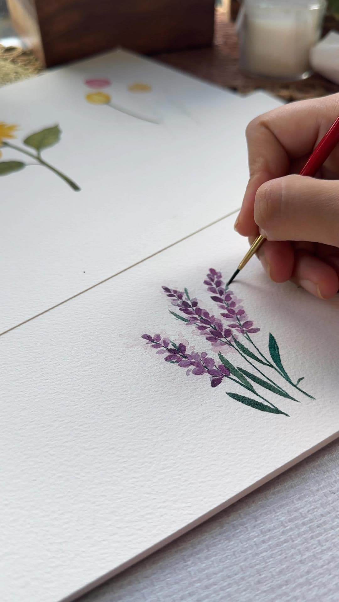 A close-up shot captures a hand delicately applying watercolor to a textured paper, painting a cluster of lavender flowers with soft purple hues and green stems. The hand, holding a fine brush, is positioned in the upper right, suggesting careful, artistic control. In the background, faint sketches of other botanical elements like yellow flowers and pink circles are visible, indicating an ongoing artistic process. The scene is set on a light-colored surface with a blurred wooden object and a glass jar in the background, creating a calm, creative atmosphere. The watercolor medium lends a translucent, painterly quality to the image, emphasizing the delicate nature of the artwork and the artist's focus. The overall mood is tranquil and meditative, evoking a sense of quiet concentration and artistic expression.