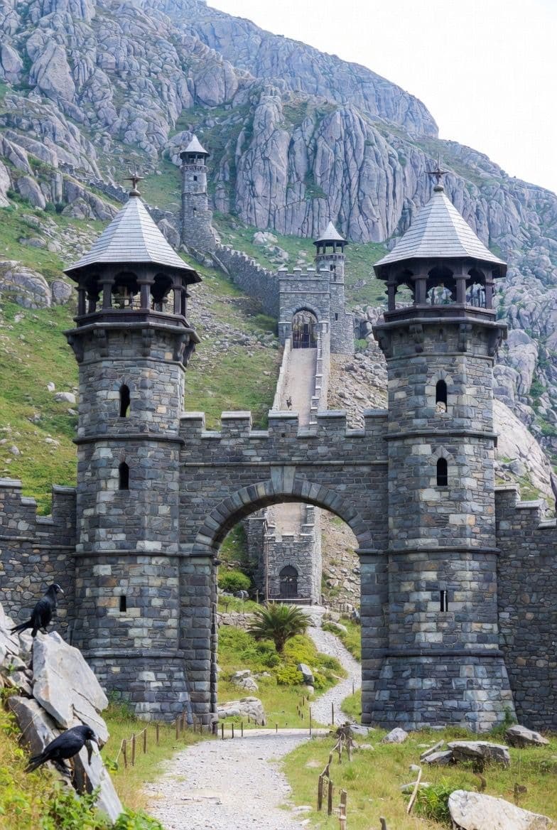 The image depicts a grand, medieval-style stone fortress gate with two towering cylindrical turrets, each topped with a conical roof and featuring arched windows. A wide stone archway leads into a narrow, winding path that ascends toward a distant castle structure nestled against a rugged, rocky mountain range. The scene is set in a natural, mountainous landscape with green grass, scattered rocks, and a few black crows perched on boulders in the foreground. The architecture is detailed with realistic stone textures and weathering, rendered in a photorealistic style. The overall atmosphere is serene and majestic, evoking a sense of ancient history and quiet grandeur under clear daylight conditions.