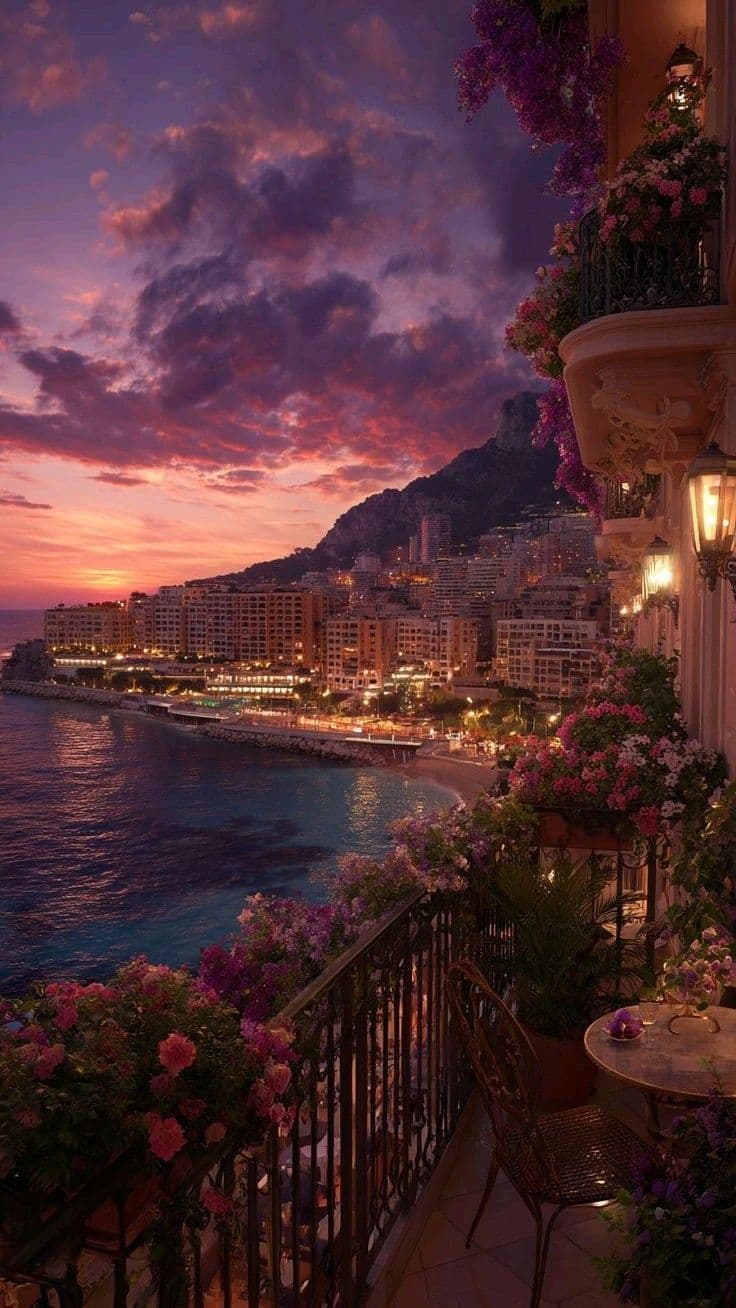 The image captures a serene coastal sunset viewed from an ornate balcony adorned with abundant flowering plants and a small table with a teacup. Below, a picturesque Mediterranean cityscape unfolds along a curving coastline, with buildings illuminated by warm lights against a dramatic sky of purples, pinks, and oranges. The scene is framed by a wrought-iron railing and a hanging lantern, evoking a tranquil, romantic atmosphere. The lighting is soft and diffused, with the sun setting behind distant hills, casting long shadows and enhancing the rich color palette. The overall composition suggests peaceful contemplation, with the viewer invited to share in the quiet beauty of the moment.