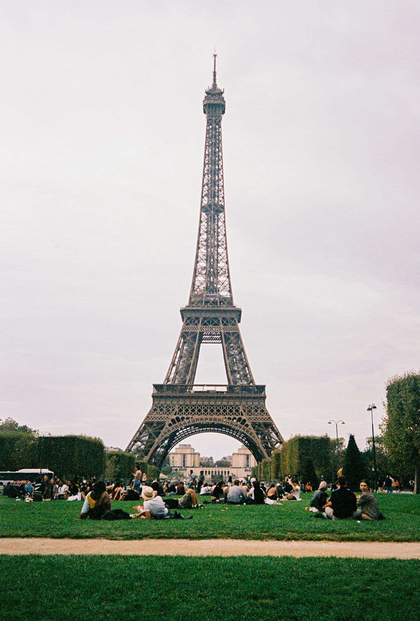 The image captures a wide-angle view of the Eiffel Tower in Paris, standing tall against a soft, overcast sky, with its intricate iron lattice structure clearly visible. In the foreground, a large grassy park area is populated by numerous people relaxing, sitting, or lying on the lawn, dressed in casual attire suitable for a public gathering. The composition emphasizes the scale of the landmark, with the tower dominating the center of the frame, while the crowd adds a sense of liveliness and human scale. The lighting is diffused daylight, creating even illumination without harsh shadows, and the color palette is muted with earthy greens and grays, enhancing the tranquil, everyday atmosphere. The overall mood is peaceful and observational, evoking a sense of leisure and cultural appreciation in a famous urban setting.