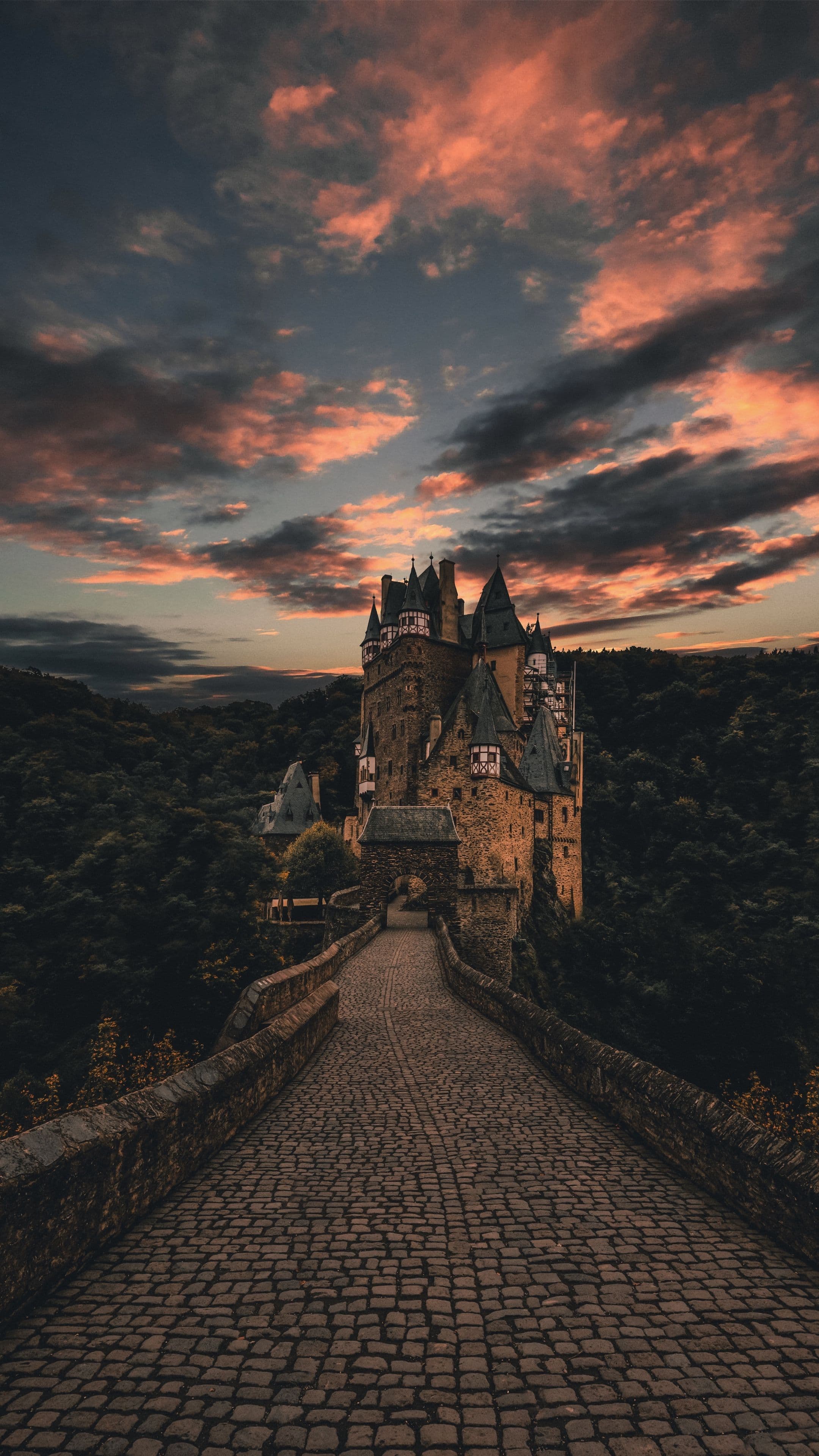 The image captures a majestic medieval castle perched atop a hill, viewed from a cobblestone pathway leading directly toward its entrance. The castle features multiple turrets, steeply pitched roofs, and stone walls, all rendered with architectural detail under a dramatic twilight sky. The sky is filled with voluminous clouds streaked with fiery orange and deep indigo hues, suggesting either sunrise or sunset. The surrounding landscape is dense with dark green forest, enhancing the castle's imposing presence. The composition uses strong linear perspective, drawing the viewer’s eye along the path to the castle’s central archway, creating a sense of journey and grandeur. The lighting is natural and atmospheric, with soft shadows and warm highlights accentuating the textures of stone and sky.