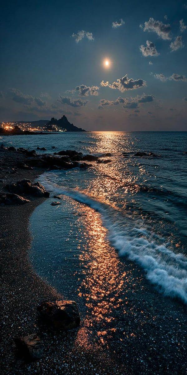 This image captures a tranquil coastal scene at twilight, featuring a luminous moon casting a golden reflection across the rippling ocean surface. Gentle waves break softly on a dark, pebbled shore, with scattered rocks adding texture to the foreground. In the distance, a silhouetted rocky headland is faintly illuminated by distant city lights, contrasting with the deep blue sky and wispy clouds. The composition emphasizes the interplay of light and shadow, with the moon’s glow creating a shimmering path on the water. The overall atmosphere is serene and contemplative, evoking a sense of peace and natural beauty through its balanced color palette and calm motion.