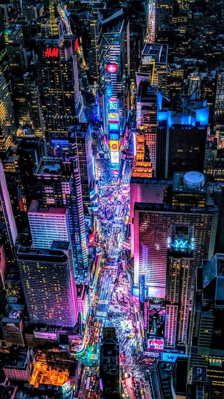 This high-resolution aerial photograph captures a vibrant, bustling urban street at night, dominated by towering skyscrapers and a dense network of illuminated billboards and neon signs. The perspective is from a high vantage point, looking down on a wide avenue filled with moving vehicles and pedestrians, creating a dynamic sense of urban energy. The lighting is predominantly artificial, with a rich palette of blues, purples, pinks, and yellows emanating from building facades and signage, contrasting with the dark sky. The composition emphasizes verticality and density, with the street receding into the distance, while the sharp focus and vivid color saturation enhance the visual impact. The overall atmosphere is energetic, modern, and immersive, evoking the pulse of a global metropolis.