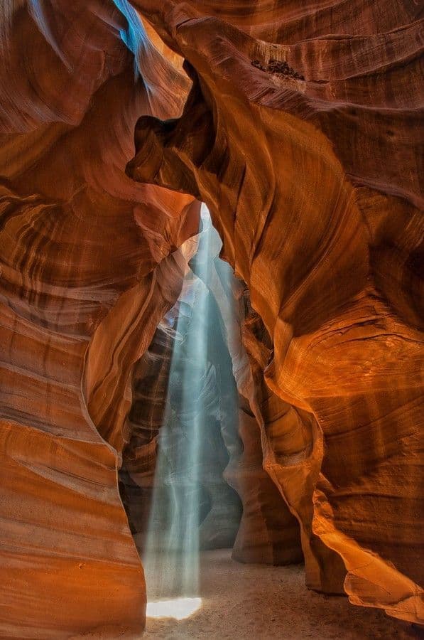 This image captures a breathtaking natural cavern with smooth, undulating sandstone walls in rich orange and red hues, sculpted by erosion over millennia. A vertical beam of cool, ethereal blue light pierces through a narrow opening at the top, illuminating the cavern floor and creating a dramatic contrast with the warm tones of the rock. The composition emphasizes the grandeur and serenity of the geological formation, with the light acting as a focal point that draws the viewer's eye into the depth of the space. The textures are highly detailed, showcasing the striations and curves of the rock surfaces, while the lighting creates soft shadows and highlights that enhance the three-dimensional form. The overall atmosphere is one of quiet awe and natural beauty, evoking a sense of peace and wonder in the viewer.
