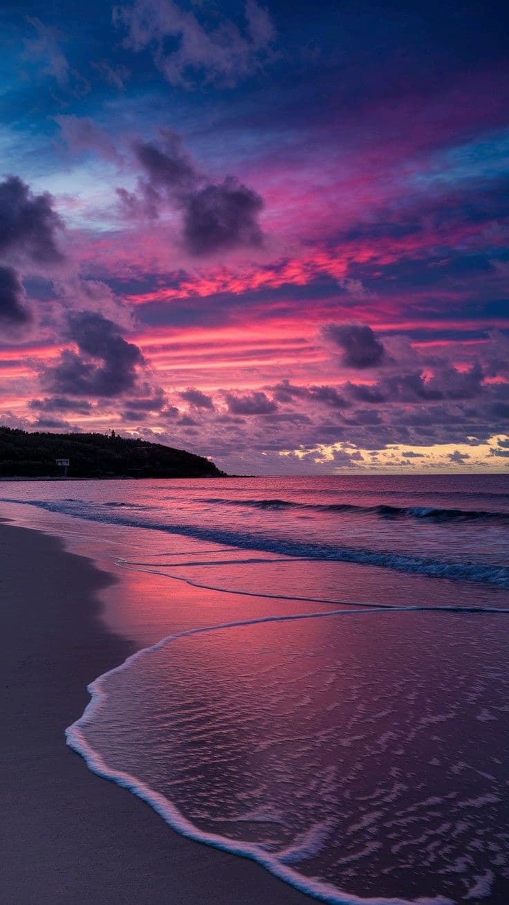 This image captures a serene coastal sunset with a dramatic, colorful sky transitioning from deep indigo to vibrant magenta and pink hues, reflected in the calm ocean waters. Gentle waves lap at the shoreline, leaving delicate foam patterns on the wet sand, while a dark, tree-lined headland anchors the left side of the frame. The composition emphasizes the vastness of the natural landscape, with the horizon dividing the sky and sea in a balanced, tranquil manner. The lighting is soft and diffused, characteristic of late afternoon or dusk, enhancing the peaceful and contemplative mood. The artistic rendering features smooth gradients and realistic textures, capturing the ephemeral beauty of the moment with high fidelity and emotional resonance.