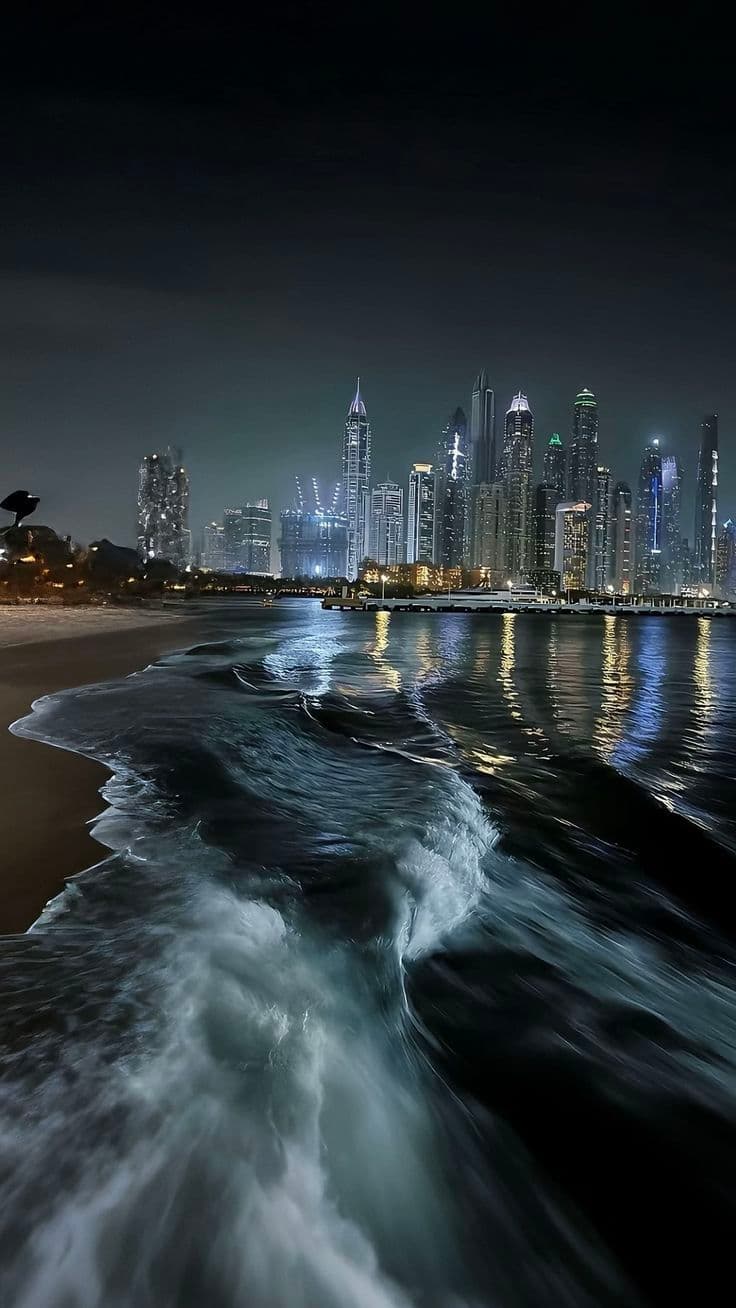 This image captures a dynamic nighttime coastal scene with a long-exposure photograph of waves crashing onto a dark shoreline, their motion blurred into smooth, white streaks. In the background, a modern urban skyline of illuminated skyscrapers reflects in the rippling water, creating a striking contrast between natural movement and architectural stillness. The composition emphasizes the interplay of light and shadow, with the city lights casting a cool, blue-toned glow over the wet sand and water. The overall atmosphere is serene yet powerful, evoking a sense of urban tranquility juxtaposed with the relentless energy of the ocean. The image is rendered with high technical precision, showcasing the interplay of motion blur, reflection, and architectural detail.