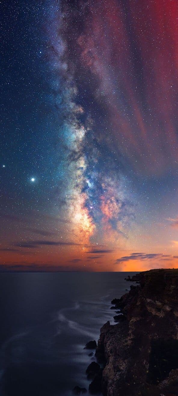 This image captures a breathtaking celestial panorama with the Milky Way arching dramatically across a twilight sky, transitioning from deep indigo to warm orange hues near the horizon. Below, a tranquil ocean meets a rugged, dark cliffside, with gentle waves lapping against the rocks in a long-exposure blur. The composition emphasizes the vastness of the cosmos juxtaposed with the earth’s serene coastline, evoking a sense of awe and solitude. The artistic rendering uses high-resolution photography with precise color gradients and natural lighting, highlighting the texture of the clouds and the reflective surface of the water. The overall mood is peaceful and contemplative, inviting viewers to reflect on the beauty of the universe and the quiet majesty of nature.