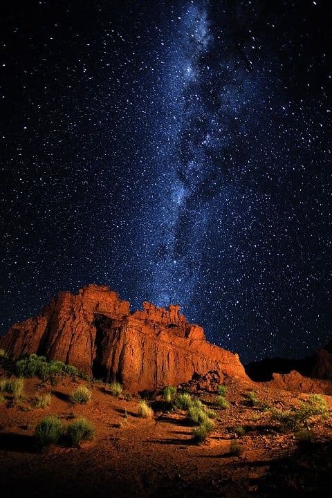 The image captures a breathtaking night sky dominated by the luminous band of the Milky Way, stretching diagonally across a dark, star-filled expanse. Below, a rugged, reddish-brown rock formation rises majestically from a sparse desert floor, its textured surface illuminated by a warm, low-angle light source that casts deep shadows and highlights its geological contours. Scattered green shrubs dot the arid terrain, contrasting with the cool blues and purples of the celestial backdrop. The composition emphasizes the vastness of the cosmos juxtaposed with the enduring solidity of the earth, evoking a sense of awe and tranquility. The photograph employs high dynamic range to preserve detail in both the bright stars and the dimly lit landscape, creating a visually immersive and emotionally serene scene.