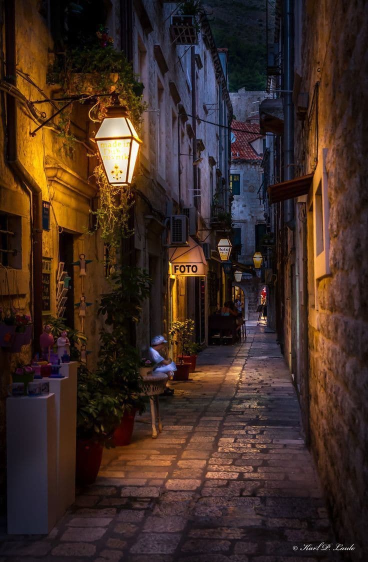 This image captures a narrow, cobblestone alleyway in a historic European town at dusk, illuminated by warm, glowing lanterns hanging from stone facades. The scene features a solitary figure seated on a bench in the foreground, wearing a light-colored hat and coat, with their back turned to the viewer, adding a sense of quiet contemplation. The architecture is characterized by aged stone walls, wrought-iron balconies, and a small 'FOTO' shop sign, suggesting a quaint, culturally rich setting. The composition uses strong leading lines created by the alley’s perspective, drawing the eye toward a distant figure in the background, enhancing depth and mystery. The lighting is soft and directional, casting long shadows and highlighting textures, while the overall atmosphere is serene, nostalgic, and inviting, evoking a sense of timeless charm.
