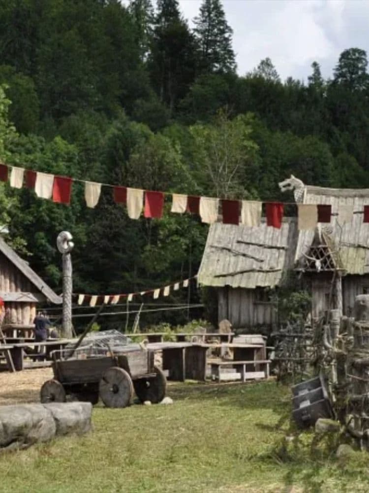 The image depicts a rustic, forest-surrounded village with weathered wooden structures and thatched roofs, evoking a medieval or Viking-inspired setting. A string of alternating red and cream-colored flags hangs across the scene, adding a festive or ceremonial touch. In the foreground, a wooden cart and stone blocks suggest a functional, lived-in environment, while a carved wooden pole and a dragon-headed roof ornament hint at cultural or mythological themes. The background is dominated by dense, green forest under a partly cloudy sky, creating a serene, natural atmosphere. The art style is photorealistic, capturing textures and lighting with high fidelity, giving the impression of a documentary-style or cinematic landscape.