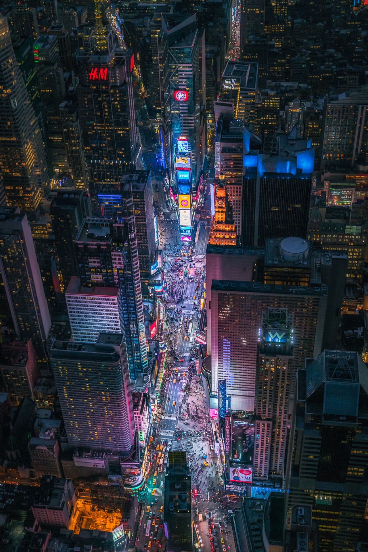 This aerial photograph captures a vibrant, bustling urban street at night, densely packed with towering skyscrapers and illuminated billboards. The perspective from above reveals a chaotic yet mesmerizing flow of traffic and pedestrians moving through the heart of a major metropolitan district. The scene is dominated by a dazzling array of neon lights and digital signage, creating a kaleidoscope of color against the dark urban skyline. The composition emphasizes the verticality of the architecture and the horizontal movement of the street below, conveying a sense of energy and modernity. The lighting is predominantly artificial, casting a warm and cool glow across the scene, enhancing the contrast between the glowing signs and the shadowed buildings.