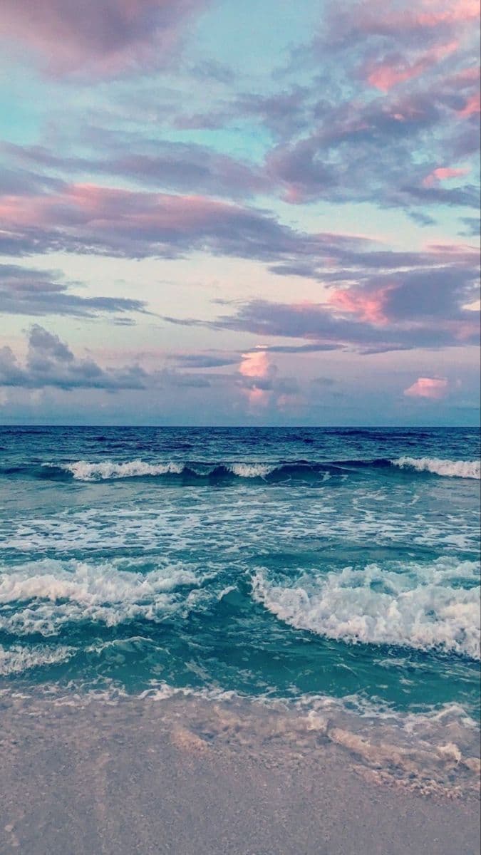 This image captures a serene coastal landscape during twilight, featuring gentle ocean waves rolling onto a sandy shore. The sky is painted with soft gradients of lavender, pink, and pale blue, suggesting either sunrise or sunset. The water displays dynamic movement with white foam cresting at the waves, while the foreground sand appears damp and textured. The composition emphasizes natural beauty and tranquility, with no human figures or clothing present. The lighting is diffused and atmospheric, enhancing the peaceful mood without any visual distractions. The overall aesthetic is calming and evocative of quiet contemplation or relaxation.