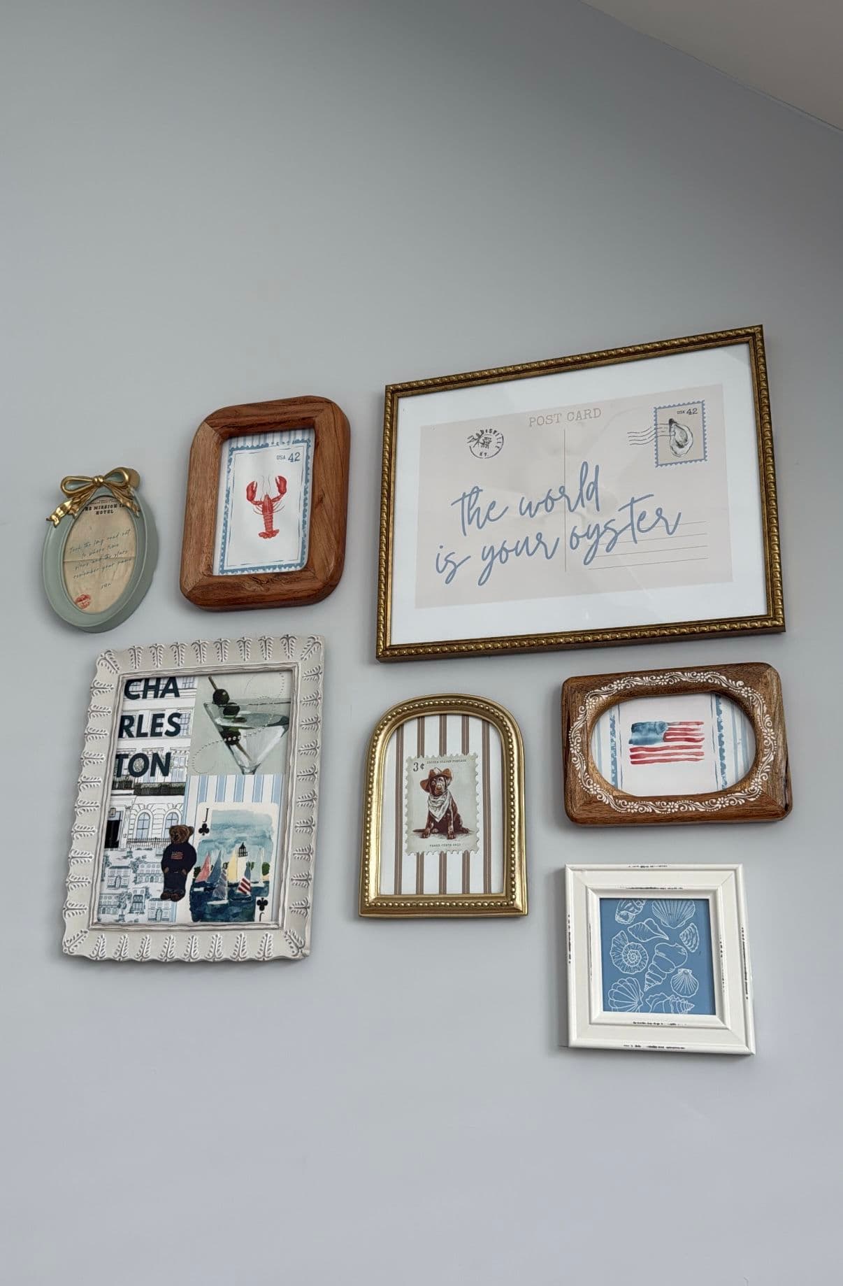 This image displays a curated gallery wall featuring six distinct framed decorative items arranged on a plain, light-gray wall. The frames vary in style, including rustic wood, ornate gold, and distressed white, each containing unique content such as a lobster illustration, a postcard with the phrase 'the world is your oyster', a collage of a cocktail and cityscape, a dog portrait, an American flag, and a blue patterned design. The arrangement is asymmetrical yet balanced, creating a visually engaging composition that suggests a personal, curated aesthetic. The lighting is even and natural, highlighting the textures and details of each frame without casting harsh shadows. The overall atmosphere is cozy, nostalgic, and inviting, evoking a sense of personal history and curated taste.