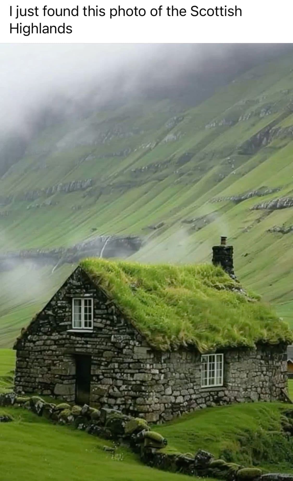 The image features a traditional stone cottage with a lush green thatched roof, nestled in a verdant, misty Highland valley. The structure is built from weathered grey stones, with white-framed windows and a dark doorway, surrounded by low stone walls and vibrant green grass. In the background, rolling green hills rise steeply under a cloudy, atmospheric sky, with a faint waterfall visible in the distance. The scene is rendered with naturalistic detail, emphasizing texture and depth, evoking a serene, timeless rural atmosphere. The lighting is soft and diffused, suggesting an overcast day, enhancing the tranquil and slightly mysterious mood of the landscape.