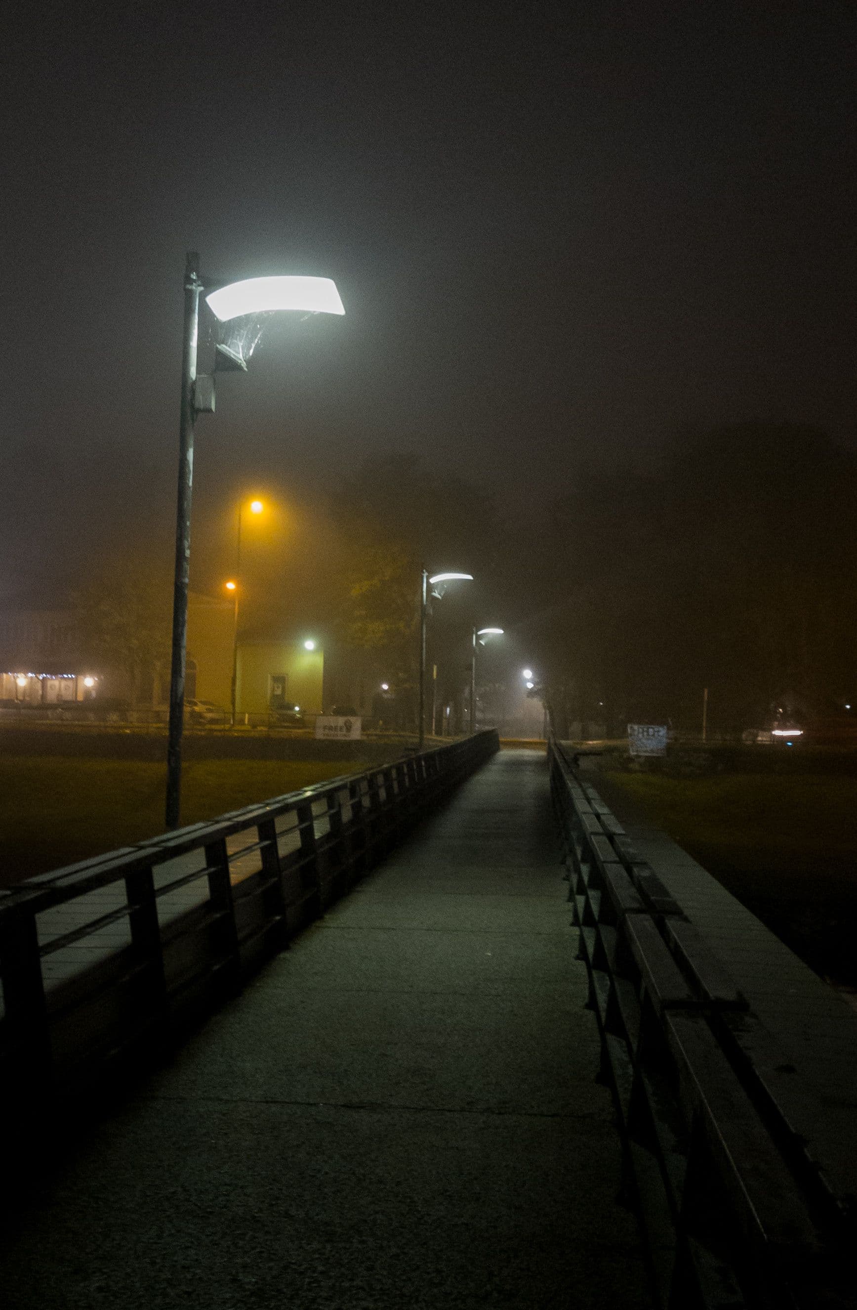 This image depicts a solitary, foggy urban pathway illuminated by streetlights, creating a moody, atmospheric scene. The perspective is from a low angle along a concrete walkway flanked by dark metal railings, leading into the misty distance. Faint yellow and white lights from distant buildings and streetlamps pierce the fog, casting soft glows against the otherwise dark, indistinct background. The fog obscures details of trees and structures, enhancing the sense of isolation and quiet mystery. The overall tone is contemplative and serene, with a muted color palette dominated by greys, blacks, and the warm glow of artificial light. The composition uses leading lines to draw the viewer’s eye into the depth of the foggy path, emphasizing solitude and stillness.