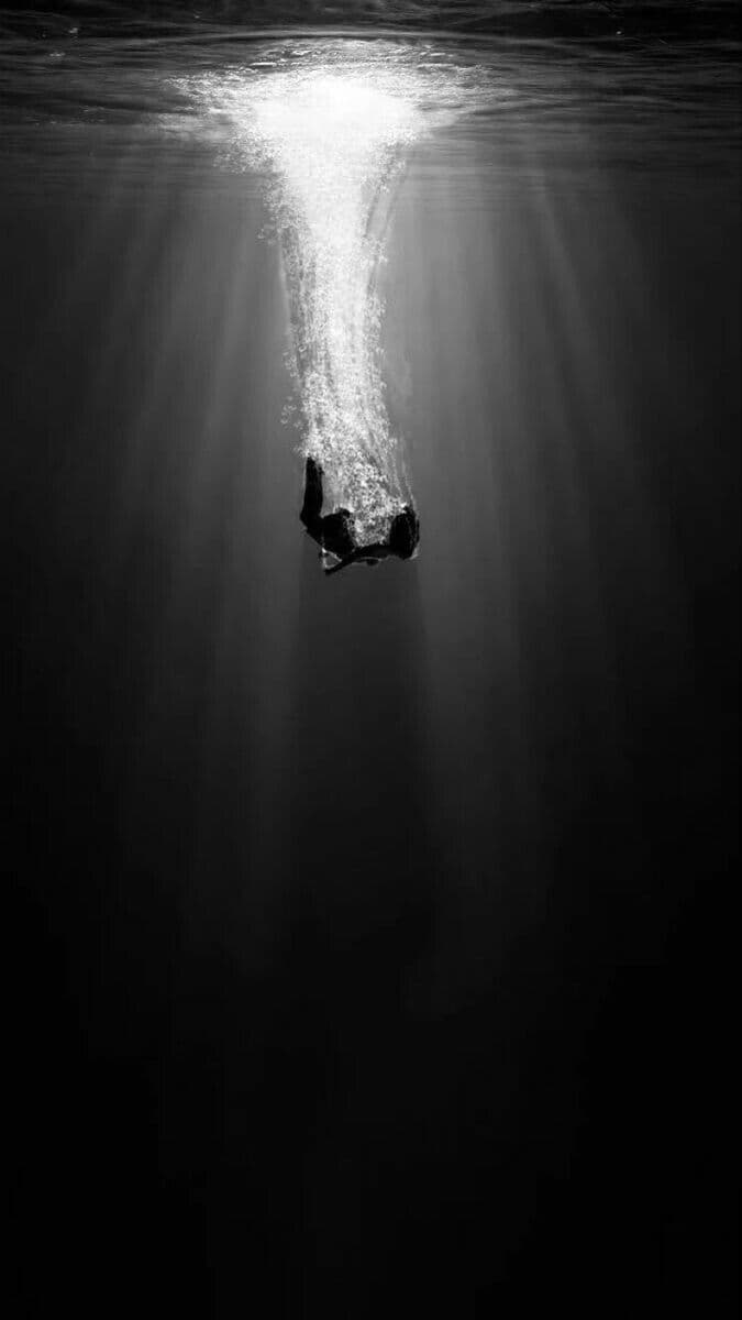 This black-and-white underwater photograph captures a solitary figure submerged in deep water, silhouetted against a brilliant beam of sunlight piercing the surface. The figure, appearing to be a human, is oriented vertically with limbs slightly bent, suggesting a slow descent or suspension. Radiant rays of light descend from the water’s surface, creating dramatic contrast and emphasizing the depth and isolation of the scene. The composition is minimalist, focusing entirely on the interplay of light, shadow, and the solitary human form. The overall mood is contemplative and serene, evoking a sense of mystery and quiet introspection through its stark, high-contrast visual language.