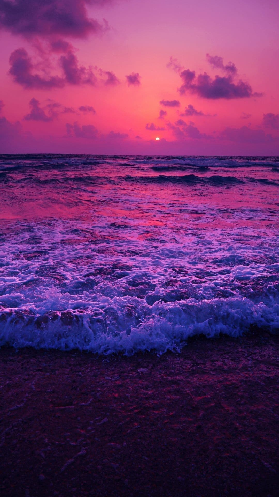 This image captures a tranquil coastal sunset with a vivid, saturated color palette dominated by deep purples and magentas. The sun, a small glowing orb, hovers just above the horizon, casting a radiant glow across the sky and reflecting onto the rippling ocean surface. Waves gently break along the shore, their white foam contrasting with the richly colored water and dark sand. The composition emphasizes the natural beauty of the seascape, with clouds softly scattered in the upper atmosphere, enhancing the serene and peaceful mood. The lighting is soft and diffused, characteristic of twilight, creating a dreamlike and calming atmosphere suitable for relaxation or contemplation.