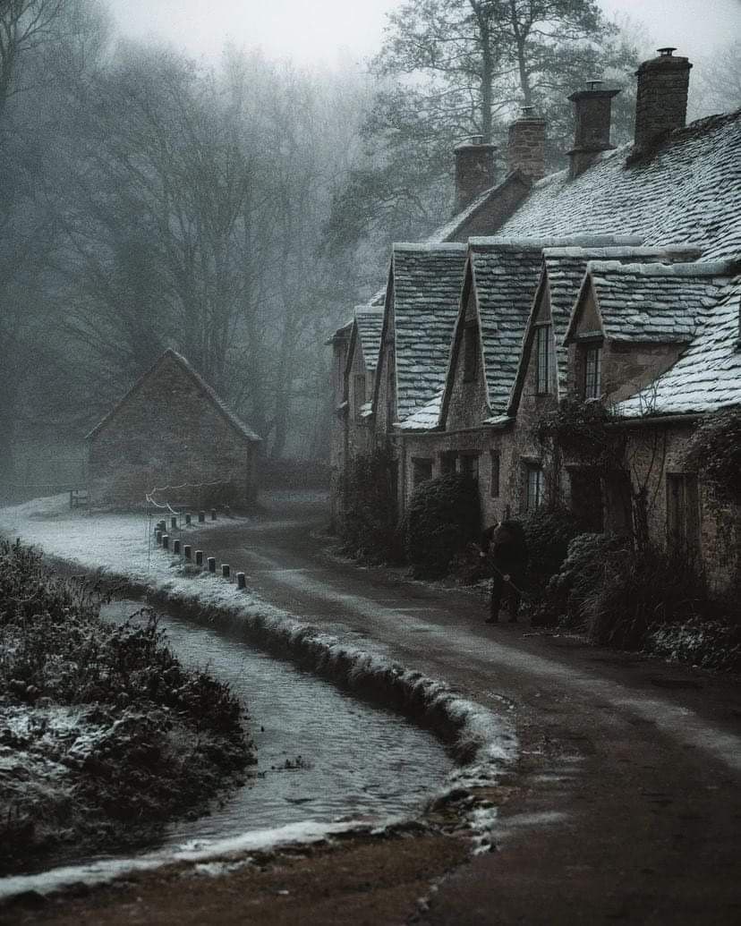 The image portrays a quiet, misty village street lined with traditional stone cottages featuring steeply pitched roofs covered in snow. A solitary figure in dark, full-length winter attire walks along the wet, snow-dusted road, adding a sense of solitude to the scene. The atmosphere is enveloped in soft fog, with bare trees silhouetted against a pale, overcast sky, enhancing the tranquil, almost melancholic mood. The photograph employs a muted color palette and shallow depth of field, focusing on the path and architecture while blurring the background to emphasize atmospheric perspective. The overall tone is peaceful and introspective, evoking a sense of quiet isolation in a rural, wintery setting.