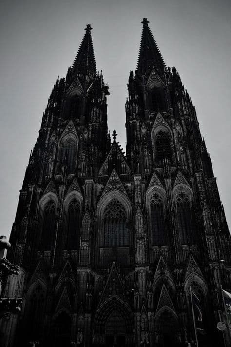 The image presents a dramatic, low-angle view of a massive Gothic cathedral with two towering spires piercing a muted, overcast sky. The architecture is richly detailed with pointed arches, ornate tracery, and vertical buttresses, emphasizing its monumental scale and historical grandeur. The monochrome palette enhances the solemn, imposing atmosphere, while the stark contrast between the dark stone and the pale sky creates a powerful visual statement. The composition is symmetrical, drawing the viewer’s eye upward along the central axis of the structure, evoking a sense of awe and reverence. The overall mood is solemn, majestic, and timeless, capturing the spiritual and architectural significance of the landmark.