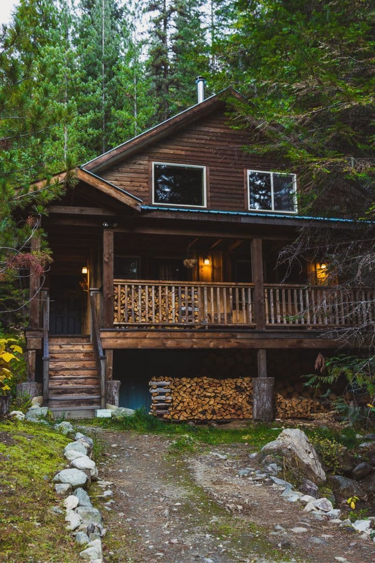 A rustic wooden cabin with a prominent front porch and stacked firewood beneath its elevated foundation, nestled deep within a dense forest of tall evergreen trees. The cabin features two windows on the upper level and warm, glowing lanterns illuminating the porch, suggesting a cozy, lived-in atmosphere. A dirt path lined with stones leads to the cabin's entrance, flanked by mossy ground and scattered rocks, enhancing the natural, secluded setting. The image employs natural daylight with soft, diffused lighting, emphasizing textures like wood grain and forest foliage. The overall mood is tranquil and inviting, evoking a sense of peace and retreat from urban life, with a strong emphasis on nature and simplicity.