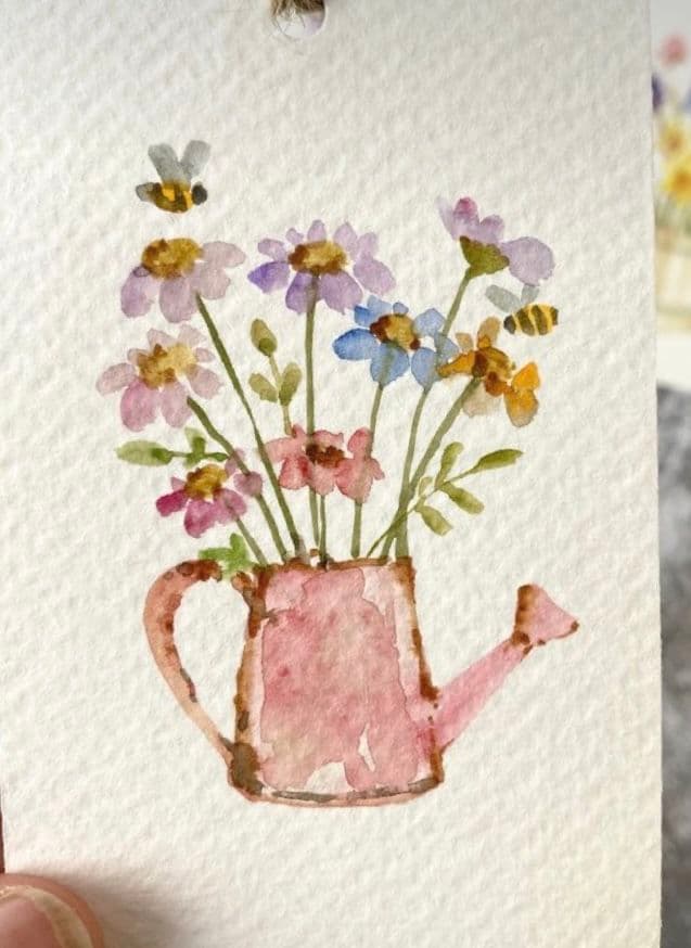 This watercolor illustration depicts a charming pink watering can overflowing with a vibrant bouquet of daisies and other wildflowers in shades of purple, blue, pink, and yellow. Two small bees are delicately rendered in flight, adding a whimsical touch to the composition. The artwork is set against a textured, off-white paper background, emphasizing the soft, painterly quality of the medium. The watercolor technique creates gentle gradients and translucent washes, giving the flowers and can a delicate, organic appearance. The overall mood is serene and pastoral, evoking a sense of gentle nature and quiet joy through its simple, handcrafted aesthetic.
