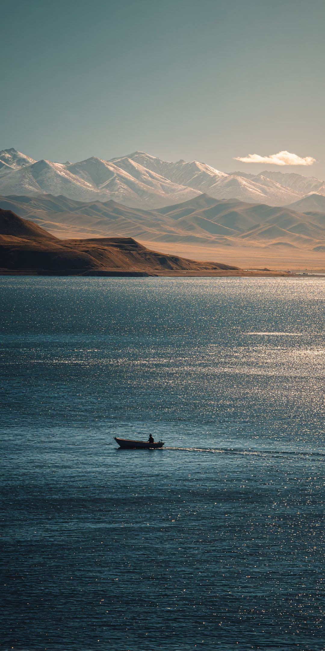 The image captures a solitary figure in a small rowboat, silhouetted against the vast expanse of a deep blue lake, gently moving across the water's surface. In the background, majestic snow-capped mountains rise in layered tiers, their peaks catching the soft light of a clear day. The foreground features rippling water with sparkling reflections, while the distant shoreline blends earthy tones with the cool blues of the lake. The composition emphasizes scale and solitude, with the lone boat serving as a focal point against the grand natural backdrop. The lighting is warm and diffused, suggesting late afternoon or early morning, enhancing the tranquil and contemplative mood of the scene.