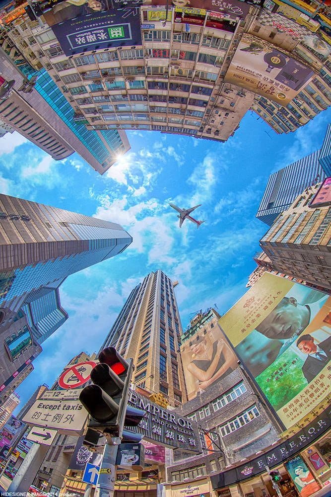 This image captures a dynamic, upward-looking perspective of a dense urban street scene, framed by towering buildings that converge toward a bright blue sky. A commercial airplane flies across the sky, adding a sense of scale and movement to the composition. The street is bustling with traffic signals, signage, and large billboards, including one featuring a woman in a modest, full-length dress. The architecture is modern and densely packed, with numerous advertisements and storefronts visible, suggesting a vibrant, high-density city environment. The photograph uses a fisheye lens to create a dramatic, immersive perspective, emphasizing the verticality of the buildings and the expansive sky. The overall atmosphere is energetic and urban, with clear daylight illuminating the scene with natural brightness and vivid color saturation.
