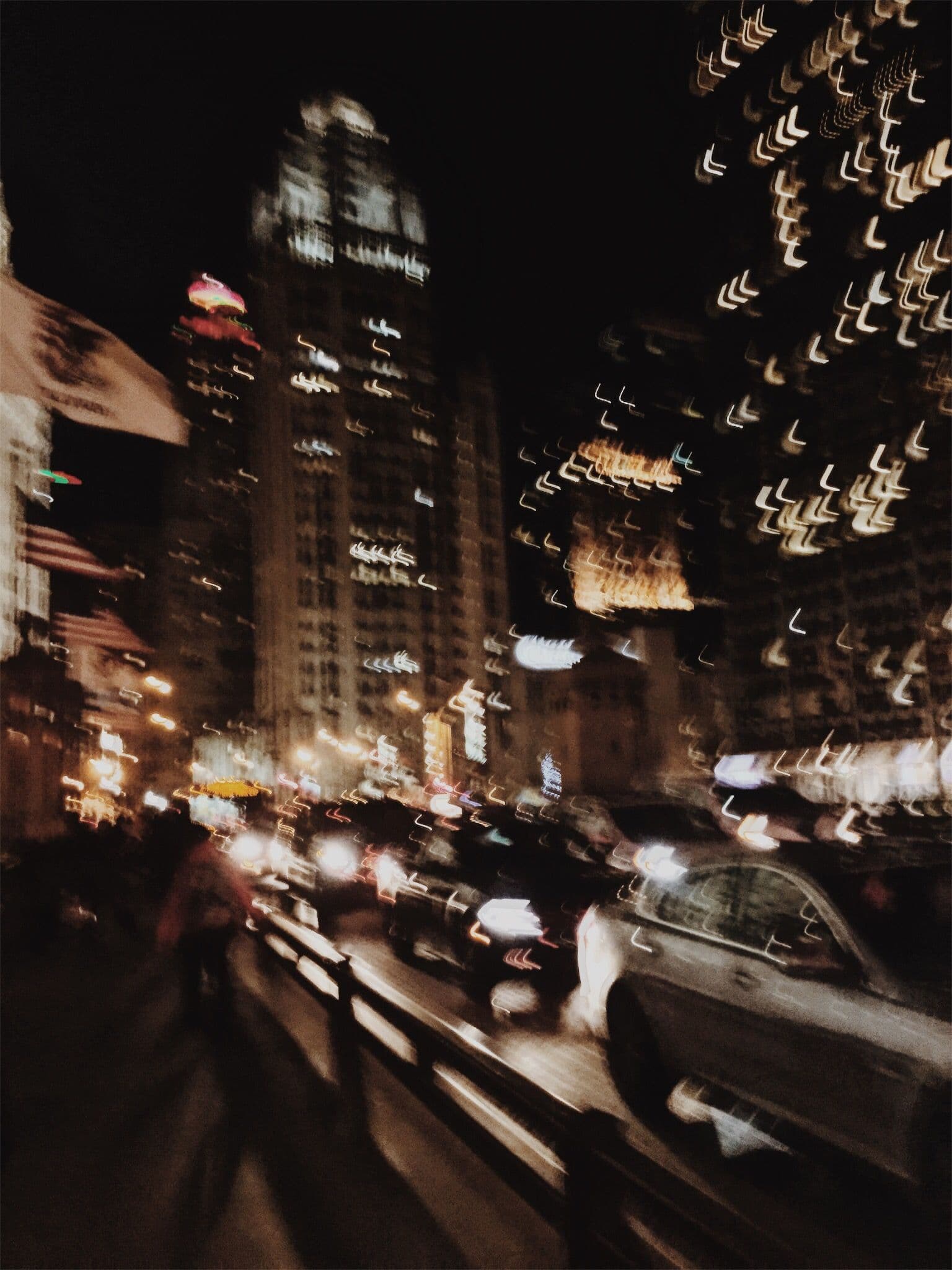 This image captures a dynamic nighttime urban street scene through a long-exposure photographic technique, creating streaks of light from vehicle headlights and city signage. The composition centers on a multi-lane road with blurred cars and pedestrians, emphasizing motion and the energy of city life. Dominant architectural structures, including a tall, illuminated skyscraper, frame the scene against a dark sky, while strings of festive lights hang overhead, adding texture and warmth. The overall aesthetic is cinematic and moody, with a shallow depth of field that isolates the foreground traffic while softly blurring the background. The color palette is dominated by warm amber and white light against deep shadows, evoking a sense of bustling urban rhythm and quiet mystery.