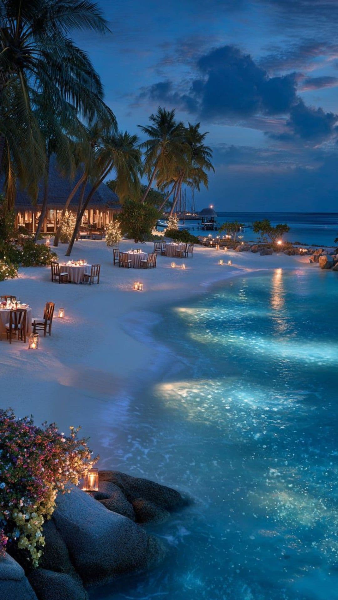 This image captures a serene, romantic beachside dining scene at twilight, featuring a luxurious infinity pool that seamlessly merges with the ocean. Soft candlelight illuminates tables set for an intimate evening, with warm glows contrasting against the cool blue tones of the water and sky. Palm trees frame the scene, adding a tropical, tranquil ambiance, while distant structures suggest a high-end resort. The composition emphasizes calmness and elegance, with gentle ripples in the water and soft lighting enhancing the peaceful, luxurious atmosphere. The overall mood is one of quiet sophistication and natural beauty, inviting relaxation and romance.