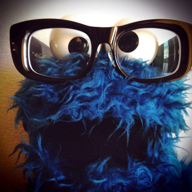 The image features a close-up, humorous depiction of the character Cookie Monster from Sesame Street. He is shown with his signature bright blue, shaggy fur and large, googly eyes, but with the addition of thick-rimmed black rectangular glasses perched on his face. The character appears to be looking directly at the camera with a wide-open mouth, creating a playful and surprised expression. The background is a simple, neutral-toned wall, which keeps the focus entirely on the puppet. The lighting is soft and diffused, highlighting the texture of the synthetic fur and the reflections in the eyeglass lenses. The overall atmosphere is whimsical, lighthearted, and comedic, subverting the character's usual wild appearance with a 'scholarly' accessory.
