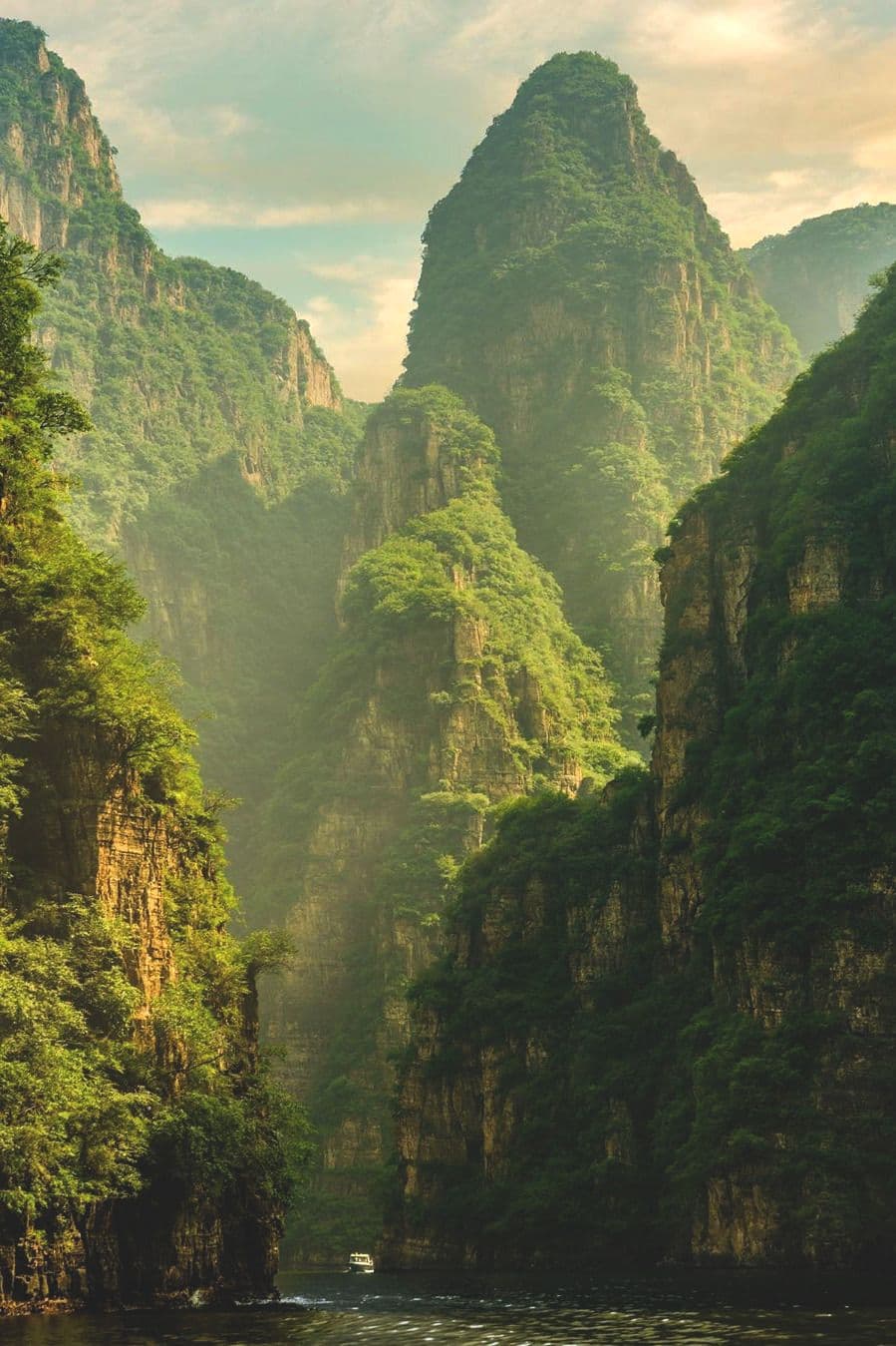 This image captures a breathtaking natural landscape featuring towering, verdant karst mountains rising steeply from a calm river. A small white boat navigates the waterway, providing a sense of scale and tranquility. The scene is bathed in soft, golden daylight filtering through a partly cloudy sky, creating a serene and majestic atmosphere. The artistic rendering emphasizes rich, layered greens and earthy tones, with realistic textures on the rock faces and foliage. The composition is balanced, with the river leading the eye toward the distant peaks, evoking a sense of peaceful exploration and awe-inspiring nature.