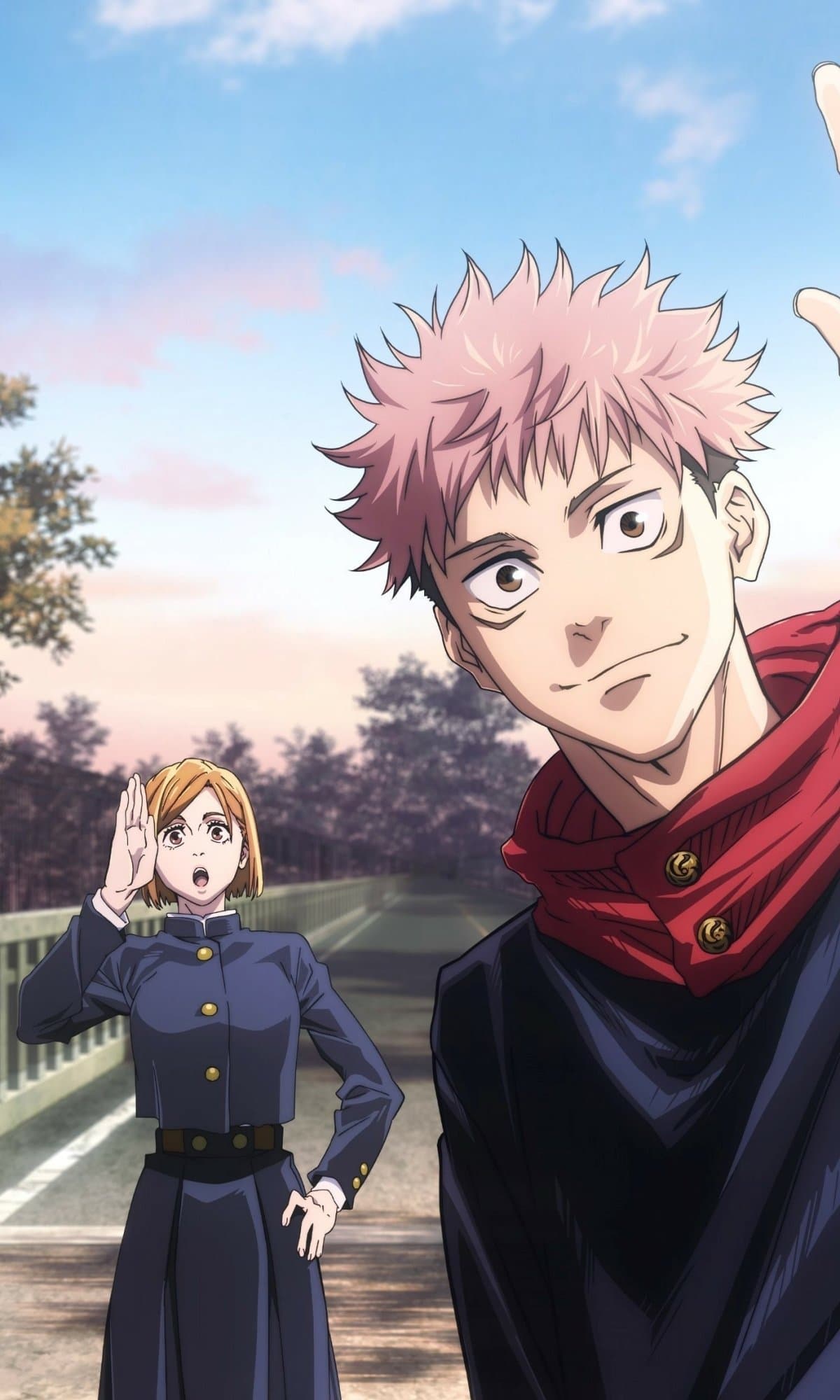 The image features two central characters, Yuji Itadori and Nobara Kugisaki, dressed in their signature dark navy high-collared school uniforms. Yuji is positioned in the foreground with a cheerful expression and spiky pink hair, while Nobara stands further back on a bridge, shouting or calling out with one hand near her mouth. The setting is an outdoor bridge or roadway during a clear day, with soft pink and blue hues in the sky suggesting early morning or late afternoon. The artistic style is characteristic of modern TV anime, utilizing sharp line work and clean cel-shading to define the characters against a slightly blurred, realistic background. The overall atmosphere is peaceful and energetic, capturing a candid, friendly moment between classmates. The composition uses a shallow depth of field to keep the focus primarily on the characters while providing a sense of place through the urban environment.